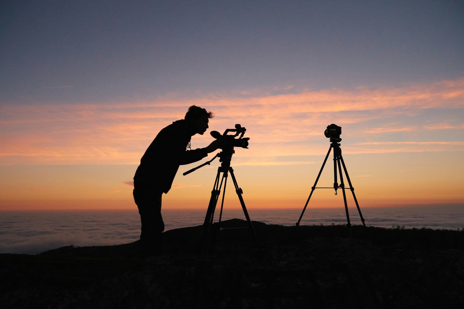 A photographer in silhouette beside a camera on a tripod on a hilltop at sunset